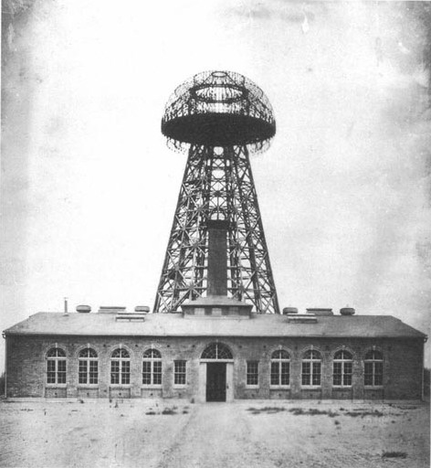 Historical photograph of Wardenclyffe Tower in 1904: a tall timber lattice tower topped by a large dome, behind Tesla's brick laboratory